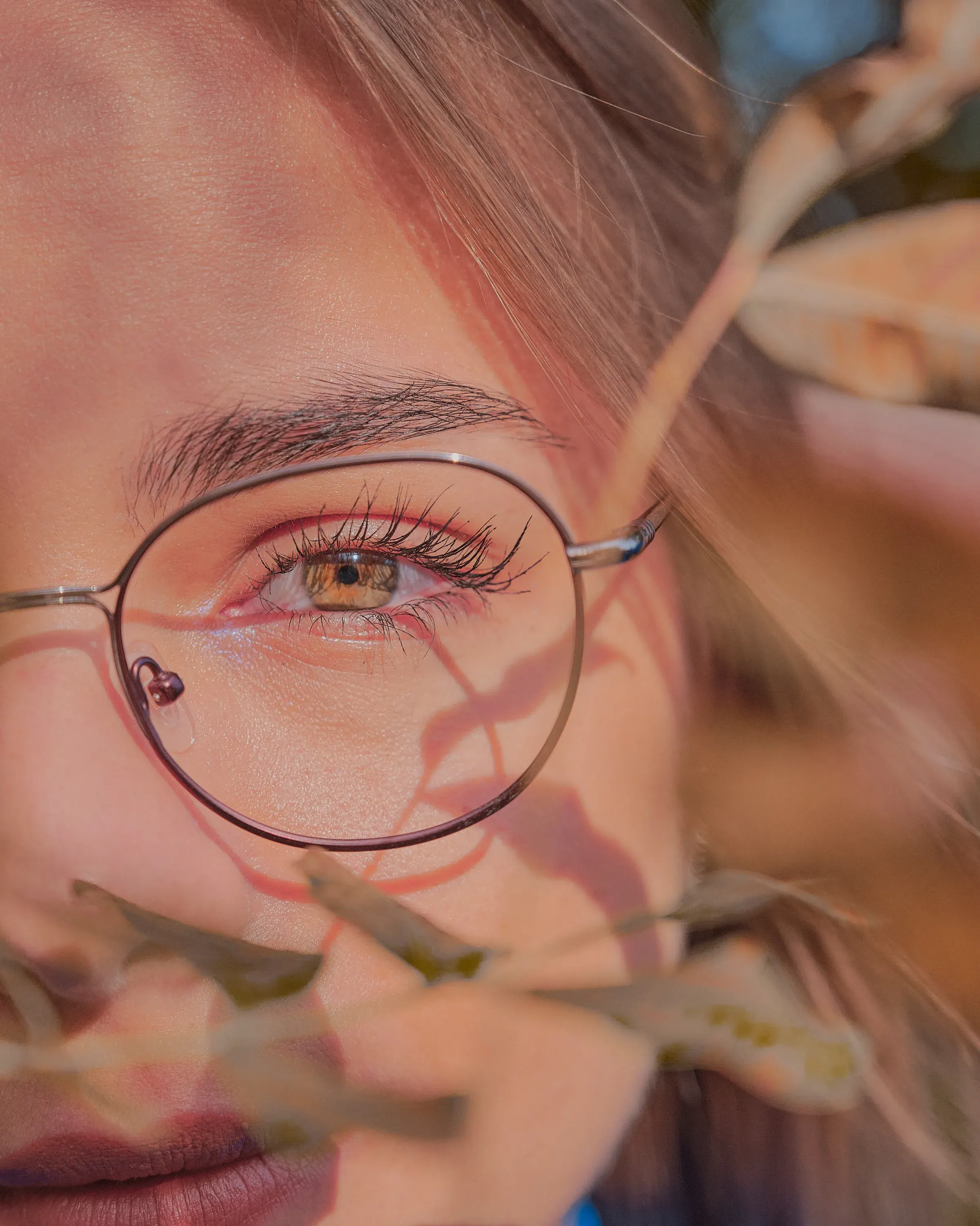 Gros plan d'un œil noisette derrière des lunettes rondes, voilé de feuillages dorés, portrait par photographe à Nice