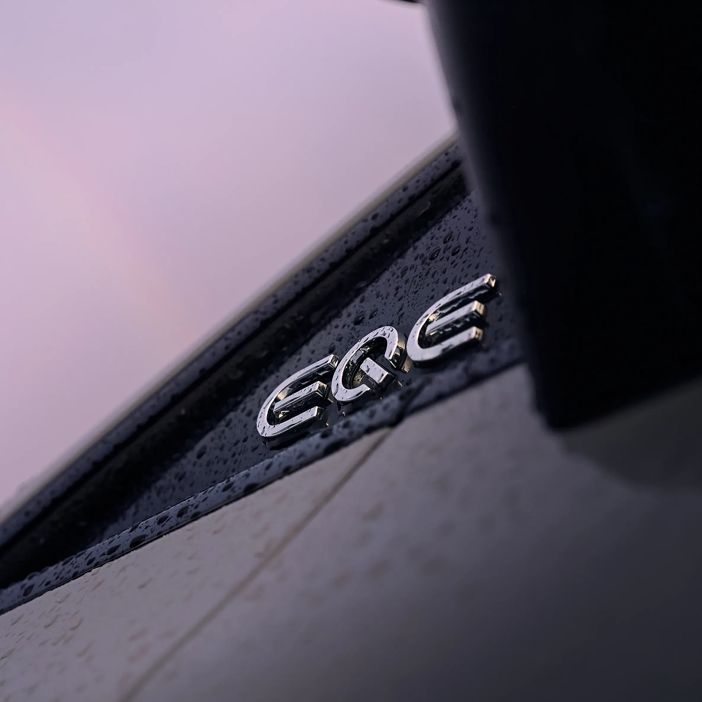 Monogramme chromé EQE perlé de pluie sur carrosserie noire, ciel mauve, photographe automobile à La Roche-sur-Foron