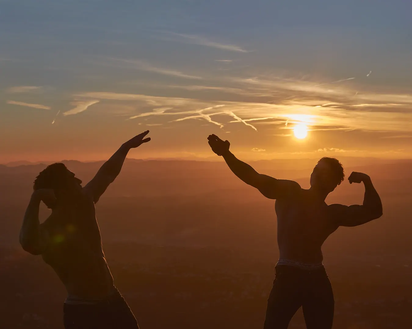 Deux athlètes torse nu en pose de musculation, silhouettes à contre-jour devant un coucher de soleil sur les collines