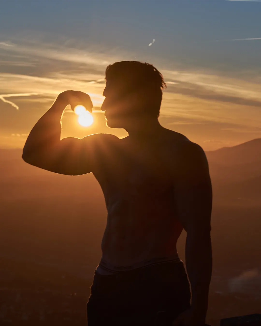 Athlète torse nu en contre-jour fléchissant son biceps face au soleil couchant sur les hauteurs, séance photo fitness