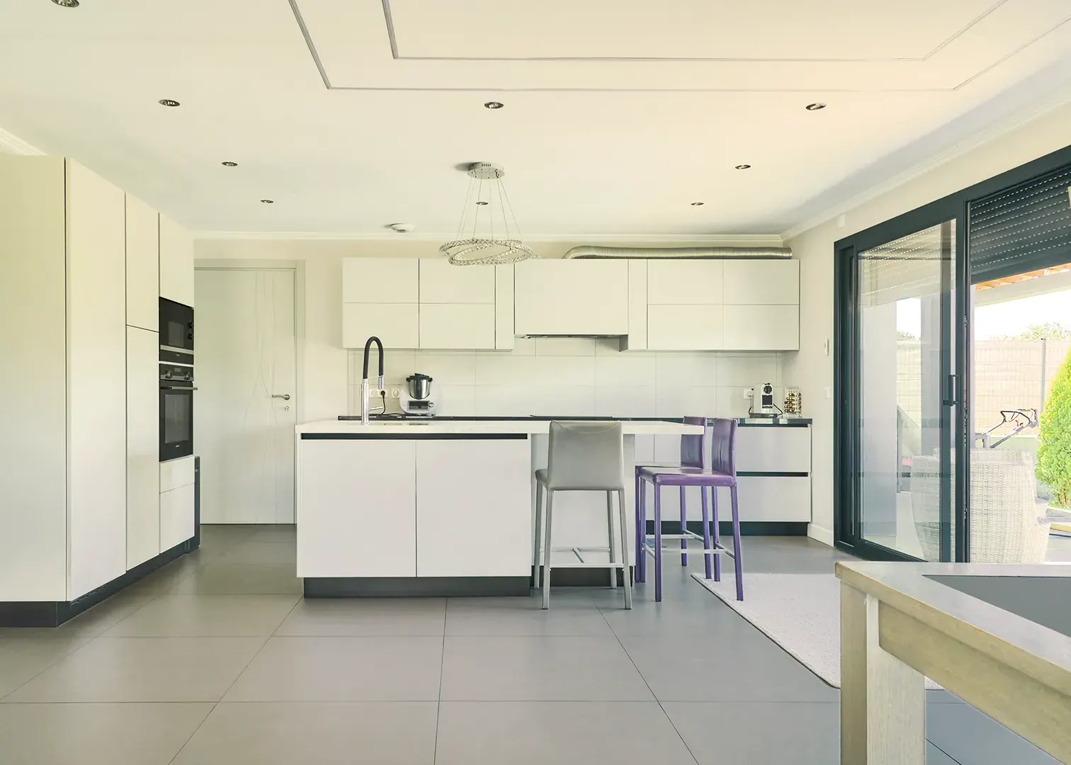 Cuisine ouverte design blanche avec îlot central et tabourets, baie vitrée sur jardin, photographie immobilier haut de gamme