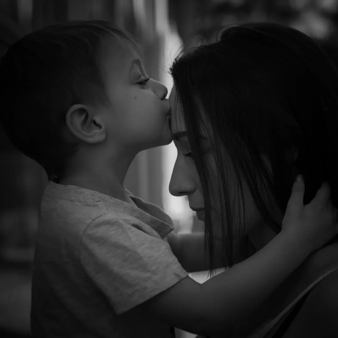 Portrait famille en noir et blanc : jeune enfant embrassant le front de sa mère, étreinte en lumière naturelle douce