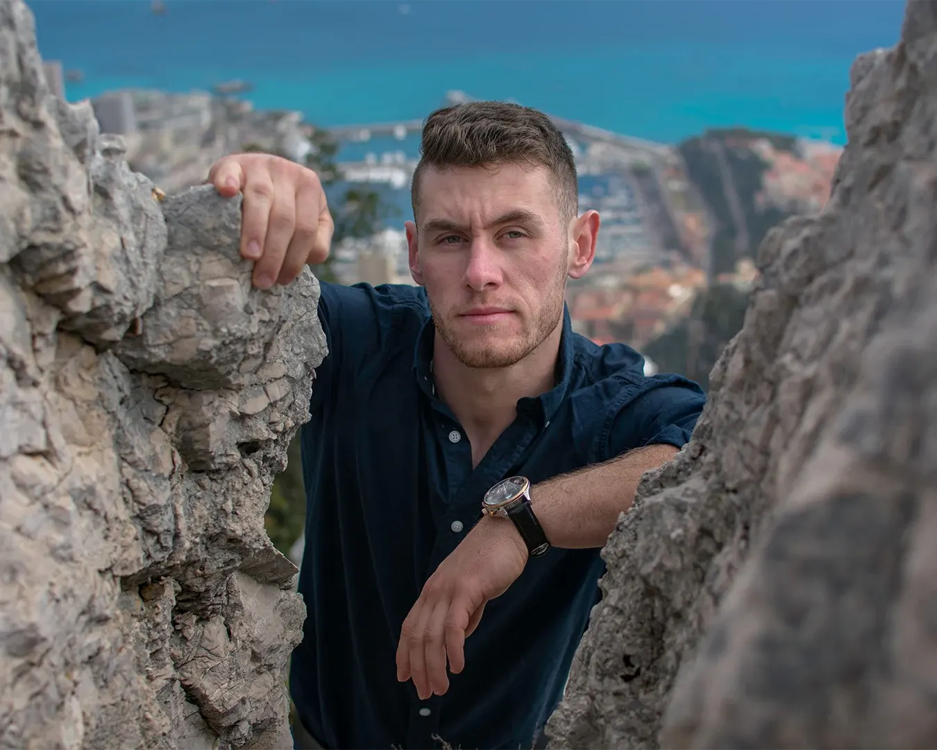 Portrait masculin en chemise bleue entre deux rochers, baie et port de la Côte d’Azur en arrière-plan