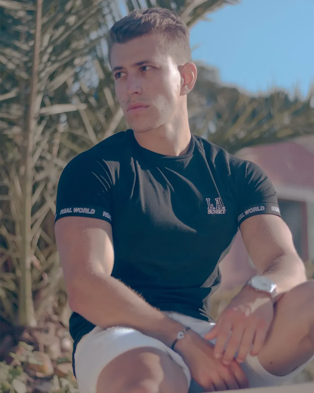 Portrait masculin en extérieur, t-shirt noir et montre, assis devant un palmier sous le soleil de la Côte d’Azur
