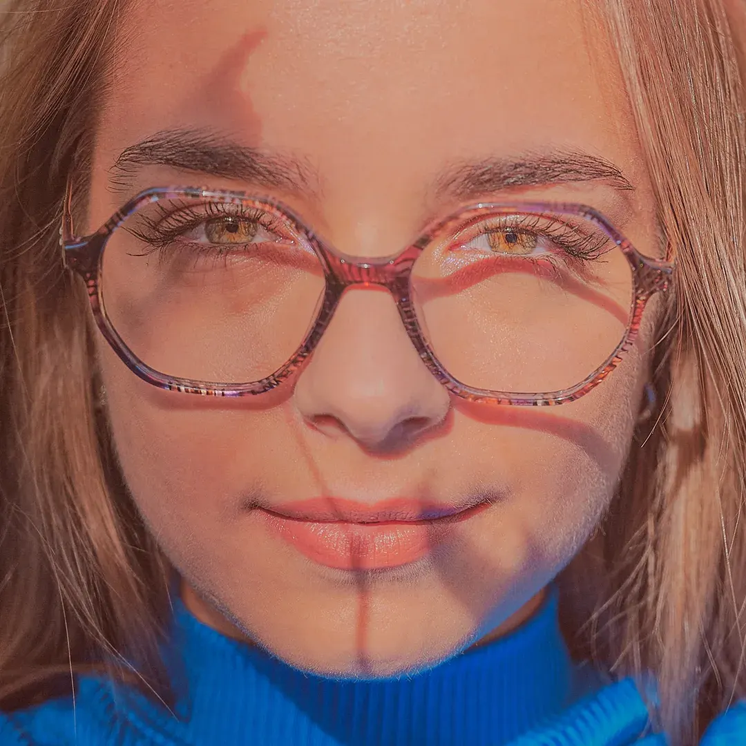 Portrait serré d’une femme à lunettes hexagonales et col roulé bleu, ombres graphiques du soleil sur le visage